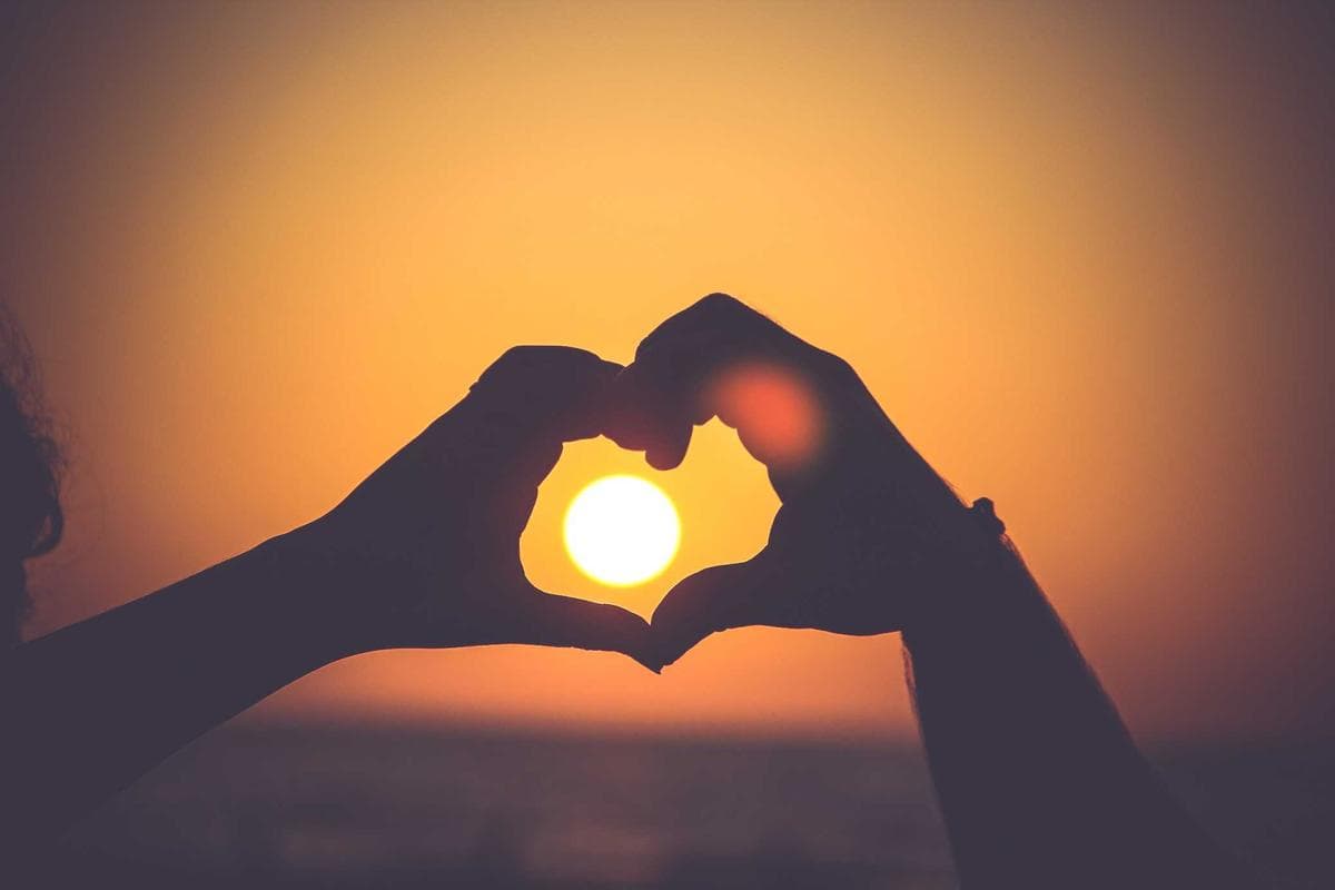 Silhouet van twee handen die een hartvorm maken tegen een fel oranje zonsondergang.