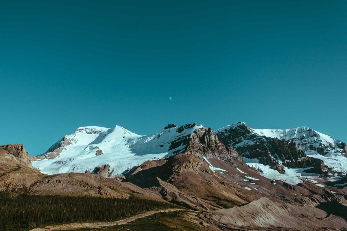 Indrukwekkend berglandschap met besneeuwde toppen en een kleine maan aan een strakblauwe hemel.