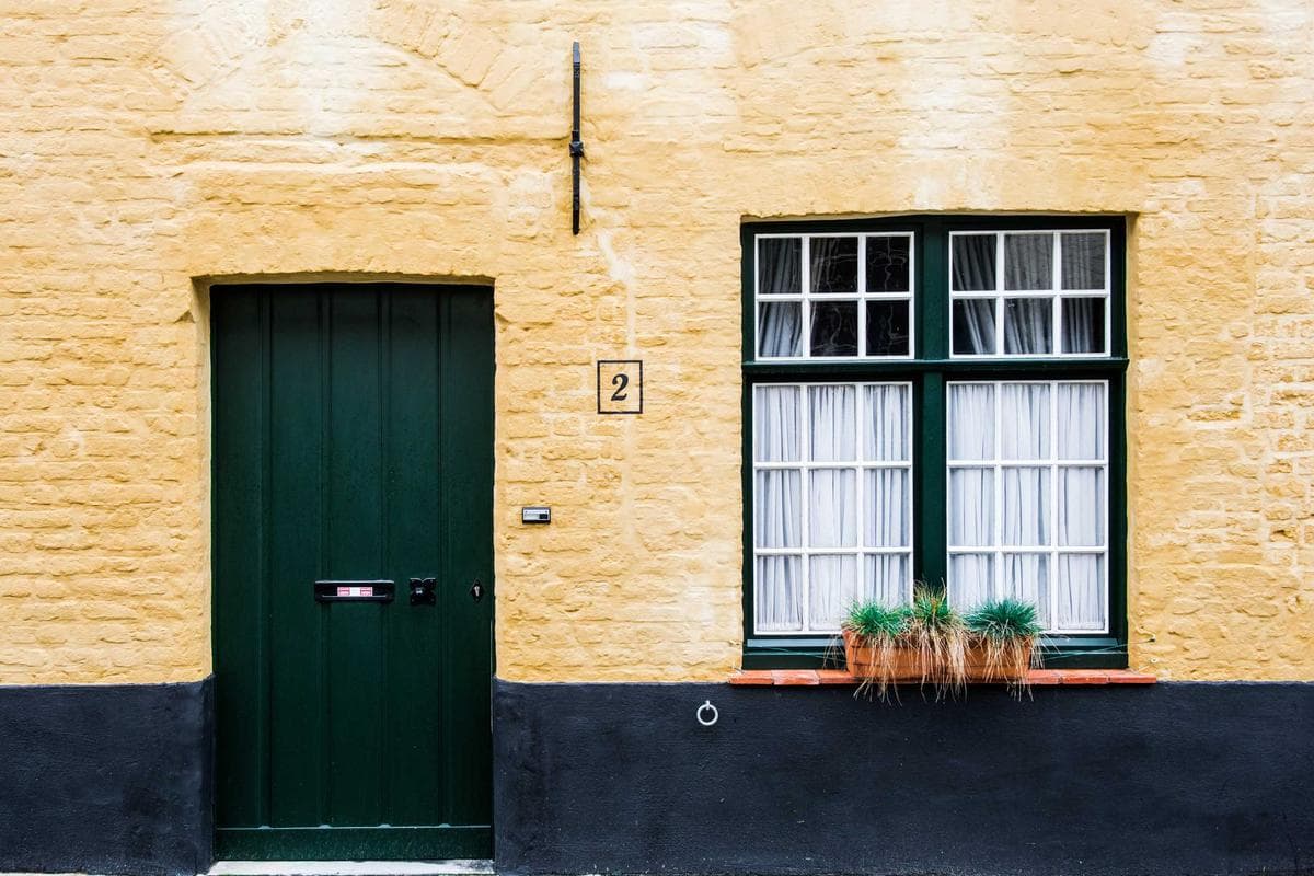 Vooraanzicht van een gele stenen muur met een groene deur, een raam met witte gordijnen en een bloembak eronder.