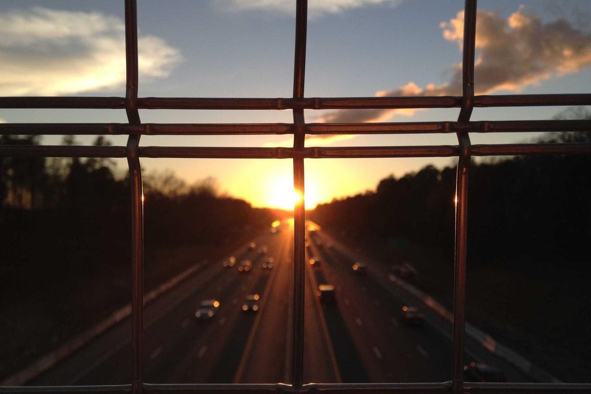 Zonsondergang boven een snelweg met rijdende auto's, gefotografeerd door de openingen van een metalen hekwerk.